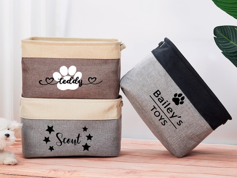 May include: Three gray and brown fabric storage bins with black paw print designs and personalized names. The bins are labeled "Teddy", "Scout", and "Bailey's Toys".