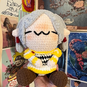 Puede incluir: Una muñeca de peluche tejida a mano con cabello de lana gris claro, cara redonda beige con una boca simple cosida en negro y orejas puntiagudas. La muñeca viste una túnica con patrón amarillo y blanco con detalles negros y pies marrones.