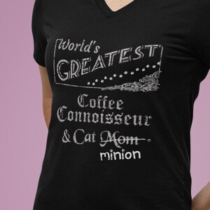 May include: Black V-neck t-shirt with white text that reads "World's Greatest Coffee Connoisseur & Cat Mom - minion." The shirt is paired with blue jeans. The text is in a decorative font.