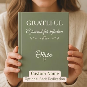 May include: A sage green hardcover journal featuring the white text GRATEFUL A journal for reflection with a decorative heart flourish. The name Olivia is printed in white script below, followed by a small heart icon. Custom name and back dedication.
