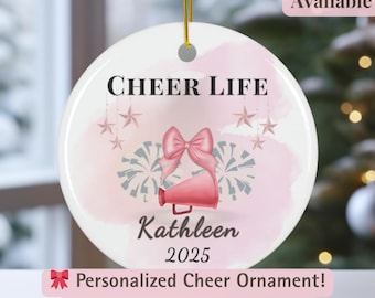 May include: A round, white ceramic ornament with the words "CHEER LIFE" in black. It features a pink megaphone, bow, and pom-poms design. The name "Kathleen" and the year "2025" are also printed on the ornament. The text "Personalized Cheer Ornament!" is also visible.
