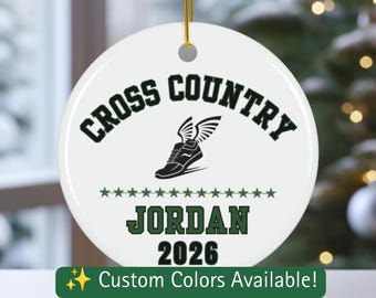 Puede incluir: Un adorno redondo de cerámica blanca con el texto CROSS COUNTRY en verde oscuro, un gráfico negro de una zapatilla de correr alada, una fila de pequeñas estrellas verdes, el nombre JORDAN en verde oscuro y el año 2026 en negro, colgado de un cordón dorado.