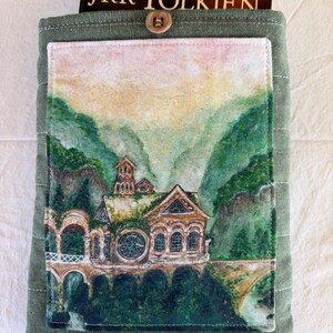 Op de afbeelding: Groene stoffen boekomslag met een knoopsluiting. De voorkant heeft een aquarel schilderij van een gebouw in een berglandschap. De boekomslag bevat een exemplaar van "The Lord of the Rings" van J.R.R. Tolkien.
