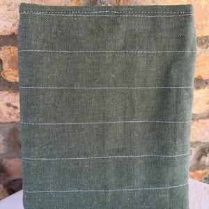 Op de afbeelding: Een rechthoekig, olijfgroen stoffen item met horizontale witte stiksels. De stof heeft een getextureerd uiterlijk. Het item staat op een wit oppervlak, met een bakstenen muur op de achtergrond.