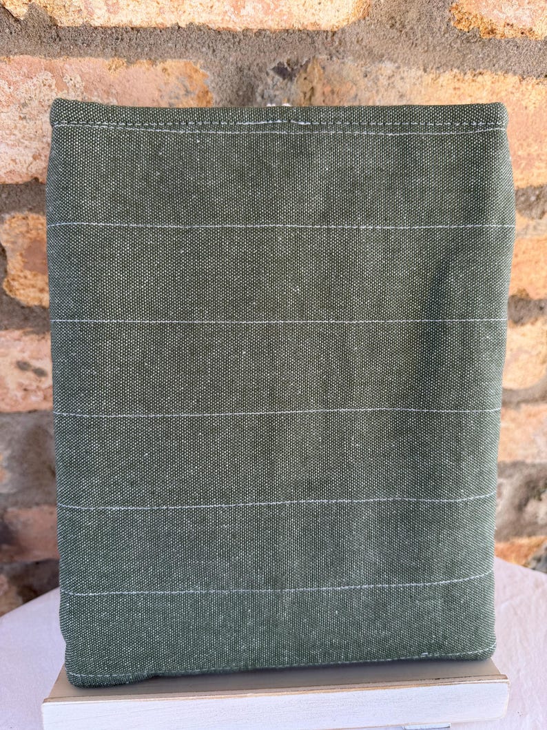 Op de afbeelding: Een rechthoekig, olijfgroen stoffen item met horizontale witte stiksels. De stof heeft een getextureerd uiterlijk. Het item staat op een wit oppervlak, met een bakstenen muur op de achtergrond.