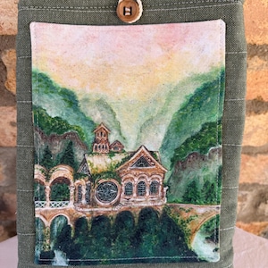 Op de afbeelding: Een groene stoffen boekomslag met een aquarel schilderij van een gebouw op een brug in een berglandschap. De hoes heeft een houten knoopsluiting en een zak. De schildering toont een gebouw met boogvensters en een groot rond raam.