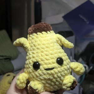 Pu&ograve; includere: Un peluche a forma di banana con corpo giallo, parte superiore marrone e piedini marroni. Il giocattolo presenta una superficie in filato lavorato all'uncinetto, due grandi occhi in plastica nera e una piccola bocca nera ricamata, formando una figura.
