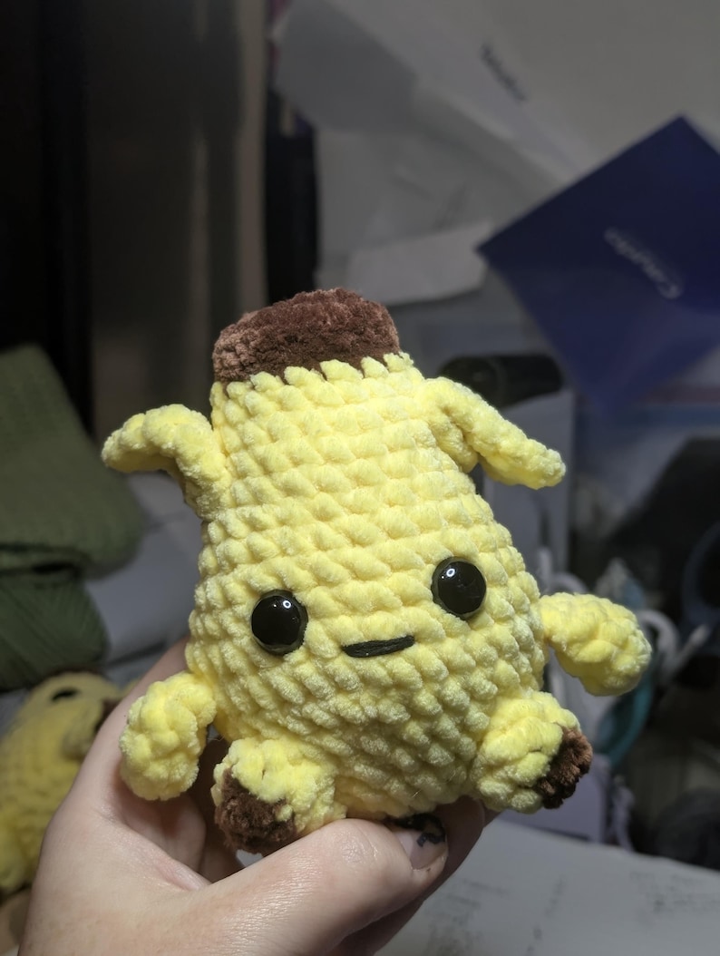 Pu&ograve; includere: Un peluche a forma di banana con corpo giallo, parte superiore marrone e piedini marroni. Il giocattolo presenta una superficie in filato lavorato all'uncinetto, due grandi occhi in plastica nera e una piccola bocca nera ricamata, formando una figura.