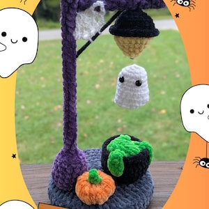 Könnte beinhalten: Handgefertigte Halloween-Dekoration aus Häkelarbeit. Sie zeigt einen lila Pfosten mit einer schwarzen Lampe, einem weißen Geist und einem Kessel mit grünem Inhalt. Ein orangefarbener Kürbis sitzt neben dem Kessel. Die Basis ist grau, und das Design ist skurril.