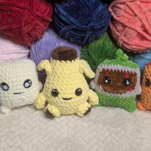May include: A collection of plush crochet toys including a white square character, a yellow banana-shaped figure with a brown top, and a green creature with a jagged mouth, all positioned in front of several large balls of colorful yarn.