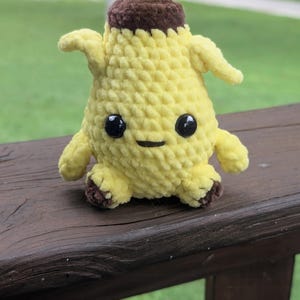 Pu&ograve; includere: Un piccolo personaggio a forma di banana realizzato all'uncinetto, con corpo giallo, parte superiore marrone e piedini marroni. Presenta due piccole braccia sui lati, due grandi occhi rotondi in plastica nera e una semplice bocca nera ricamata.