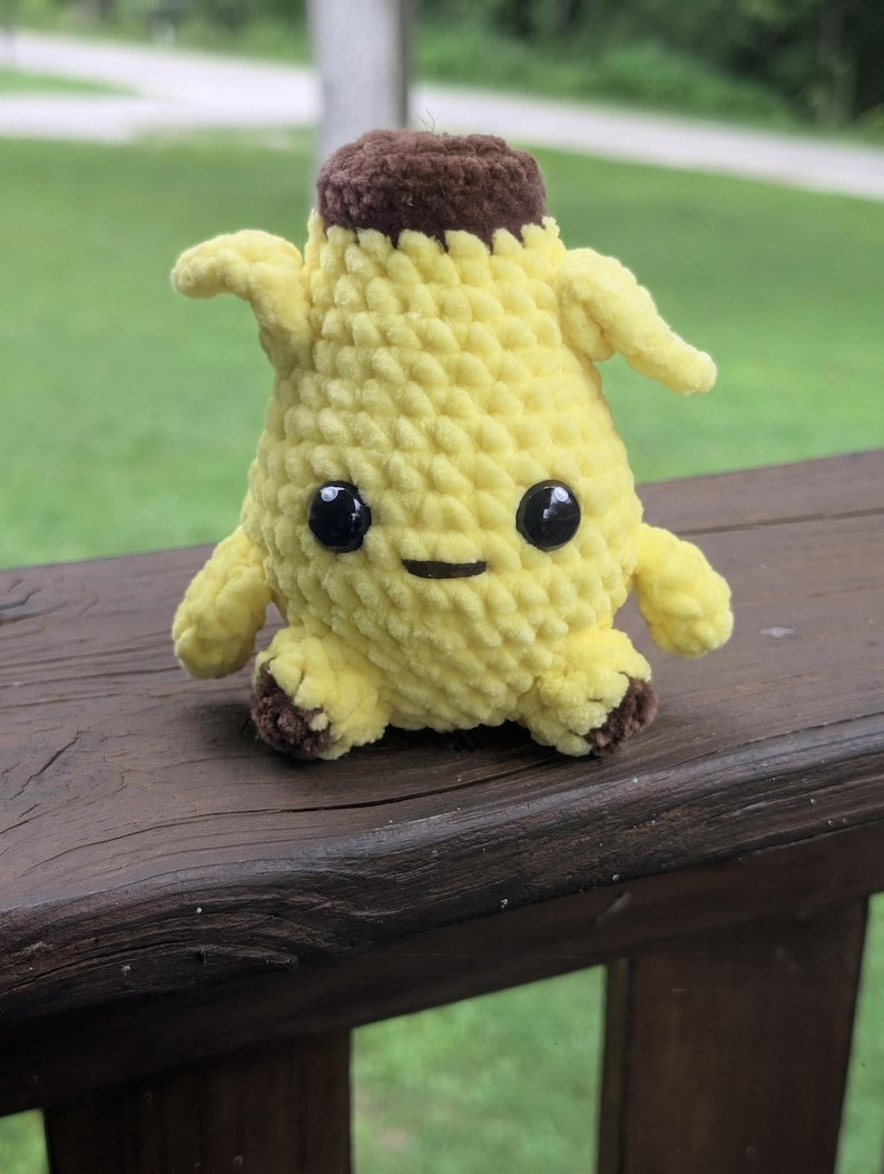 Pu&ograve; includere: Un piccolo personaggio a forma di banana realizzato all'uncinetto, con corpo giallo, parte superiore marrone e piedini marroni. Presenta due piccole braccia sui lati, due grandi occhi rotondi in plastica nera e una semplice bocca nera ricamata.