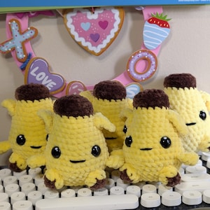 Pu&ograve; includere: Un gruppo di cinque piccoli peluche realizzati a mano all'uncinetto a forma di banana. Ogni peluche presenta un corpo giallo brillante, una parte superiore e inferiore marrone scuro, due occhi di sicurezza in plastica nera e una bocca ricamata.