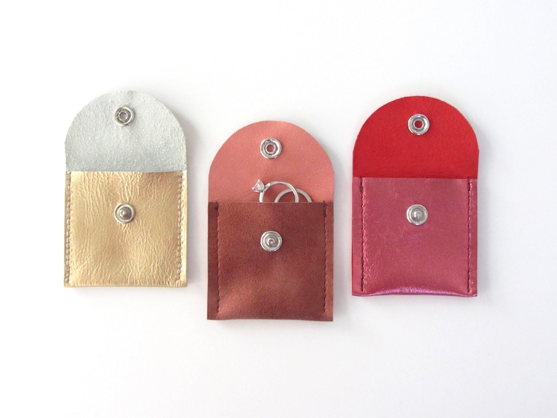 May include: Three small, square, fabric pouches with snap closures. The pouches are in gold, rose gold, and pink. The rose gold pouch has two silver rings inside.