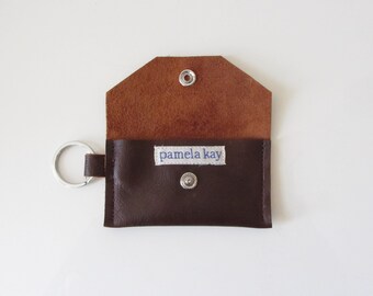 May include: A brown leather key chain with a small pocket and a silver key ring. The pocket has a white label with the text "pamela kay" printed in blue.