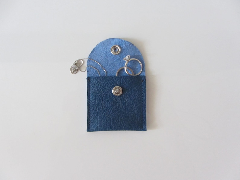 Puede incluir: Una peque&ntilde;a bolsa de cuero azul con cierre de presi&oacute;n, abierta para revelar un anillo y pendientes de plata en el interior.