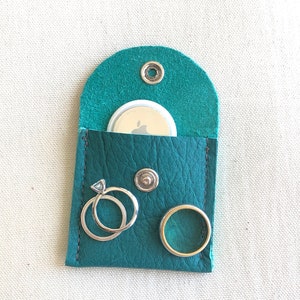 Puede incluir: Una bolsa de cuero color turquesa con solapa, un peque&ntilde;o c&iacute;rculo blanco con el logotipo de Apple, dos anillos plateados y un bot&oacute;n plateado.