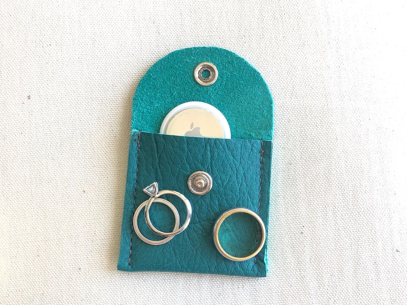 Puede incluir: Una bolsa de cuero color turquesa con solapa, un peque&ntilde;o c&iacute;rculo blanco con el logotipo de Apple, dos anillos plateados y un bot&oacute;n plateado.
