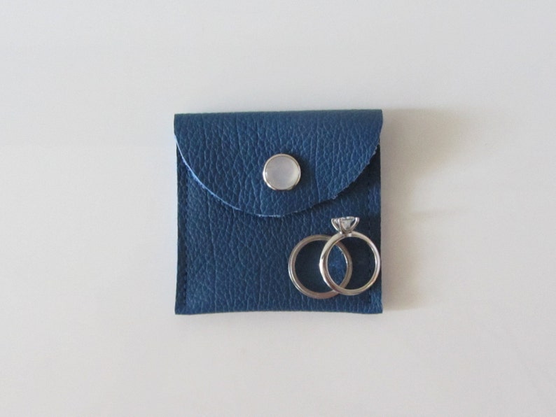 Puede incluir: Una peque&ntilde;a caja de anillos de cuero azul con cierre de bot&oacute;n blanco. Dos anillos de plata est&aacute;n dentro de la caja.