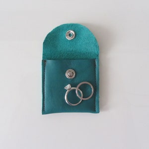 Puede incluir: Caja de anillos de cuero turquesa con dos anillos de plata en su interior. La caja tiene un cierre de presi&oacute;n.