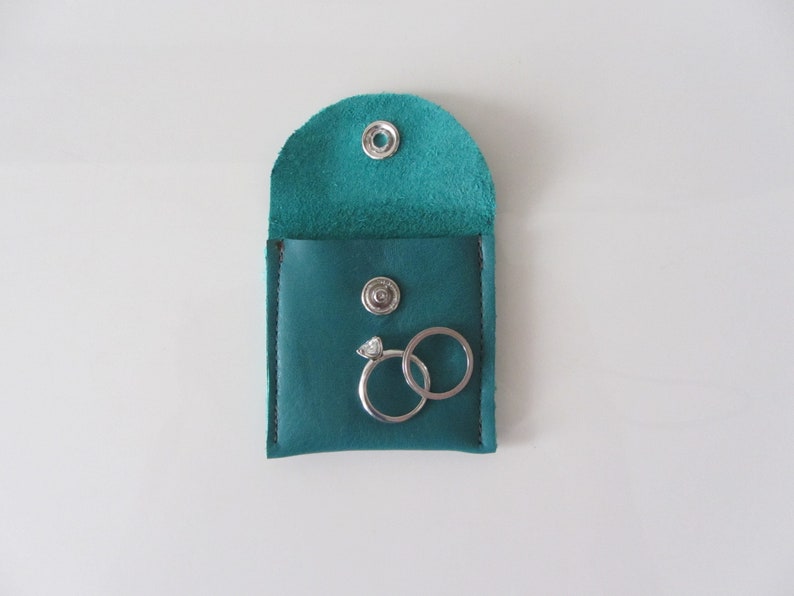 Puede incluir: Caja de anillos de cuero turquesa con dos anillos de plata en su interior. La caja tiene un cierre de presi&oacute;n.