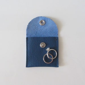 Puede incluir: Una peque&ntilde;a bolsa de cuero azul con dos anillos de plata en su interior. La bolsa tiene un cierre de presi&oacute;n y est&aacute; forrada con tela azul claro.
