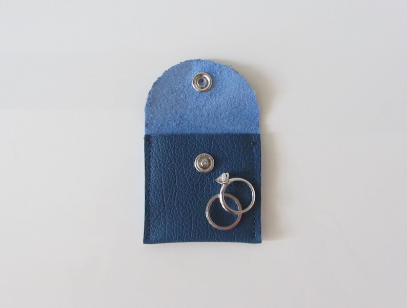 Puede incluir: Una peque&ntilde;a bolsa de cuero azul con dos anillos de plata en su interior. La bolsa tiene un cierre de presi&oacute;n y est&aacute; forrada con tela azul claro.