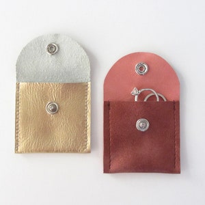 May include: Two small, square, leather pouches with snap closures. One pouch is gold with a white lining and the other is brown with a pink lining. The brown pouch has two silver rings inside.