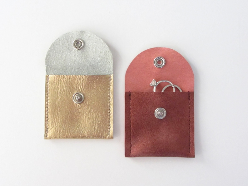 May include: Two small, square, leather pouches with snap closures. One pouch is gold with a white lining and the other is brown with a pink lining. The brown pouch has two silver rings inside.