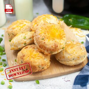 May include: A close-up of a stack of six golden brown cheddar and chive biscuits on a wooden cutting board. The biscuits are topped with shredded cheese and green onions. The biscuits are surrounded by green onions and shredded cheese.