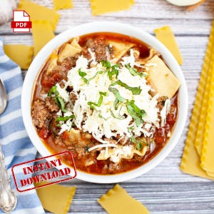 May include: A white bowl filled with lasagna soup topped with shredded cheese and fresh basil. The soup is a red color and contains noodles, ground beef, and vegetables. The bowl is sitting on a wooden table with a blue and white striped napkin and some lasagna noodles.