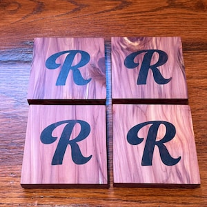 May include: Four square wooden coasters with a natural wood grain pattern. Each coaster features a large, stylized letter "R" in a dark blue color. The coasters are arranged in a square formation.