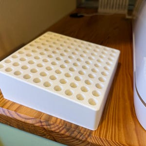 May include: White plastic storage organizer with a grid of small round holes. The organizer is sitting on a wooden surface.