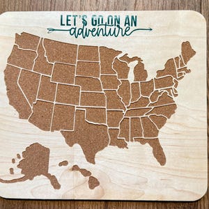 May include: A wooden map of the United States with a corkboard surface. The map is outlined in white and includes the states and their borders. The text "LET'S GO ON AN adventure" is printed in green on the top of the map.