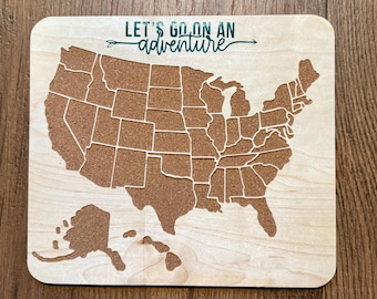 May include: A wooden map of the United States with a corkboard surface. The map is outlined in white and includes the states and their borders. The text "LET'S GO ON AN adventure" is printed in green on the top of the map.
