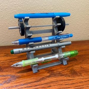 May include: A gray, 3D-printed pen holder with four pens. The pens are blue, silver, and green. The holder has a barbell design with two black circular weights. The pens are held horizontally.