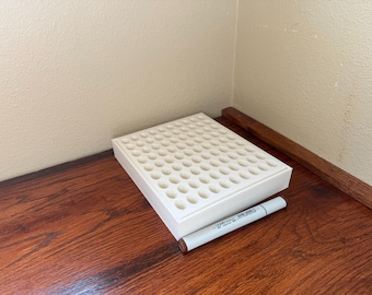 May include: A white marker storage tray with numerous circular holes, next to a gray and brown marker. The tray sits on a dark wooden surface, with a light-colored wall in the background. The marker has text on it.