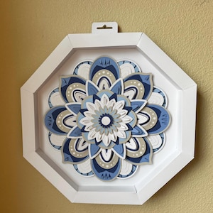 May include: A layered paper mandala art piece in a white octagonal frame. The mandala features a floral design with shades of blue, white, and tan. It has a hook for hanging. The artwork is a decorative wall hanging.