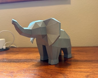 May include: A gray, geometric paper elephant sculpture with a trunk raised. The origami-style animal has a faceted design and is positioned on a wooden surface. The elephant is a decorative item.
