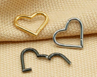 Pode incluir: Três argolas em forma de coração em metal dourado, prateado e preto. As argolas estão sobre um fundo de tecido bege.