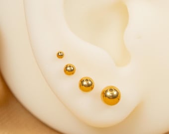 May include: Five gold-coloured ball earrings of different sizes, displayed on a white ear model.