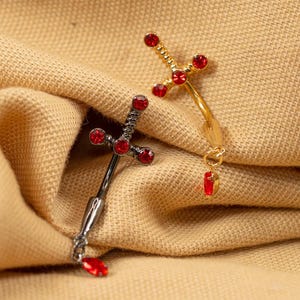 May include: Two decorative sword-shaped body jewelry pieces. One is silver-toned with red gemstones, and the other is gold-toned with red gemstones. Both have a dangling red gemstone at the bottom. The jewelry is displayed on a tan fabric.