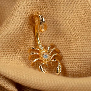 May include: Gold body jewelry featuring a scorpion design with a clear crystal in the center.