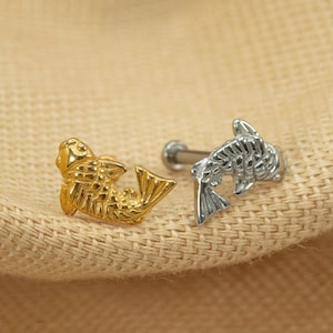 Op de afbeelding: Twee lichaamssieraden in de vorm van vissen, een in goud en een in zilver. De sieraden zijn bedoeld om in een piercing te worden gedragen.