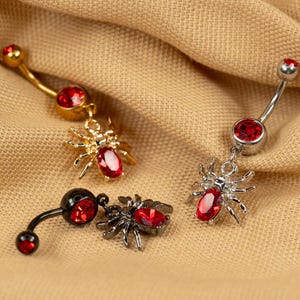 Op de afbeelding: Drie navelringen met spinnencharms. Elke ring heeft een rode edelsteen in het midden van de spin. De spinnen zijn gemaakt van goud, zilver en zwart metaal.