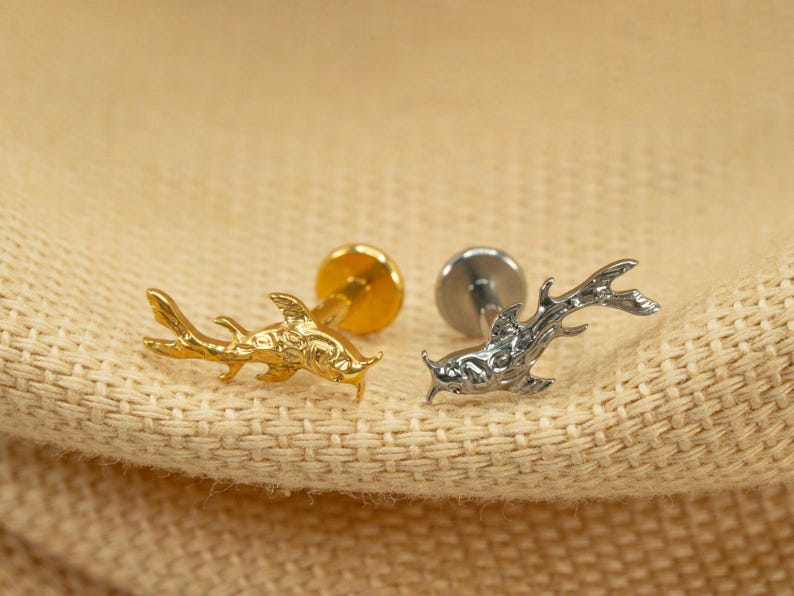 Op de afbeelding: Twee visvormige lichaamsieraden, een gouden en een zilveren, op een beige stoffen achtergrond.