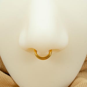 以下が含まれることがあります： ダブルフープのデザインの金の鼻のリング。リングは白いマネキンの頭につけられています。