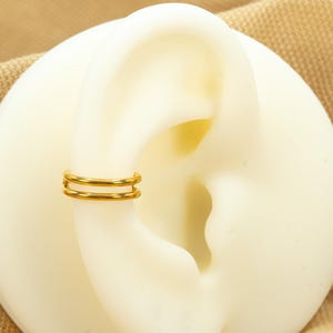 以下が含まれることがあります： 白い耳の模型につけられた、ゴールドのダブルフープイヤリング。イヤリングはシンプルで、2つのフープで構成されています。