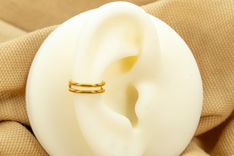 以下が含まれることがあります： 白い耳の模型につけられた、ゴールドのダブルフープイヤリング。イヤリングはシンプルで、2つのフープで構成されています。