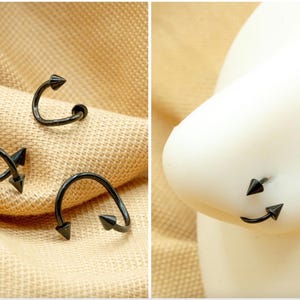 May include: Three black nose rings with pointed ends. The rings are shown on a beige fabric background and on a model's nose.